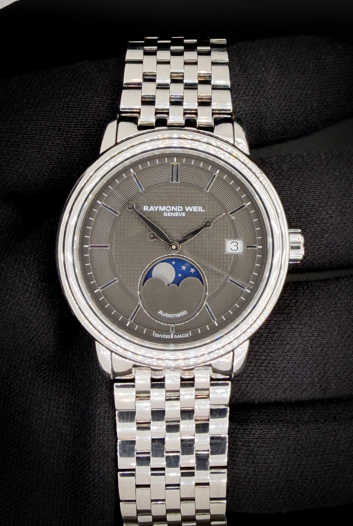 The Raymond Weil Maestro Moonphase 2879 is a classically styled dress watch that pairs traditional complications with a modern grey dial aesthetic. The polished stainless steel case measures approximately 39.5mm, offering balanced proportions that wear comfortably while maintaining an elegant wrist presence. The sunray grey dial features Roman numerals, blued-style hands, and a moonphase display at 6 o’clock, complemented by a date subdial that adds symmetry without cluttering the layout.