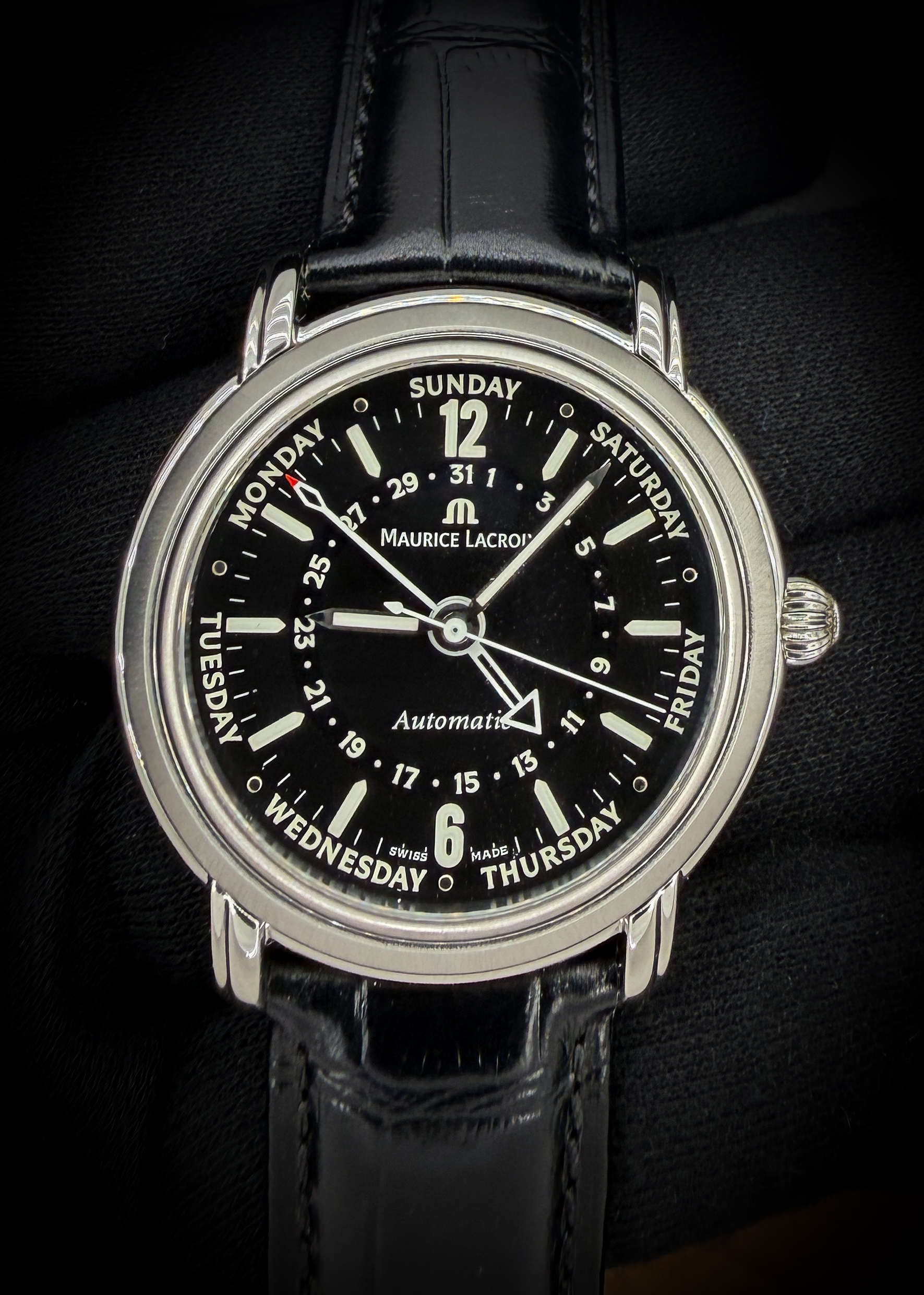 The Maurice Lacroix Masterpiece Cinq-Aiguilles MP6328 combines classic Swiss craftsmanship with a distinctive five-hand display. Its 40mm stainless steel case frames a black dial with luminous hands and hour markers, bold Arabic numerals at twelve and six, and dedicated central pointers indicating the date and day of the week. Named “Cinq-Aiguilles,” French for “five hands,” the design presents hours, minutes, seconds, day, and date entirely through centrally mounted hands. Power comes from the decorated, 25-jewel ML27 automatic movement, visible through the exhibition case back, while a sapphire crystal protects the dial. This example is fitted with a new black leather strap paired with its original Maurice Lacroix buckle, completing its elegant and individual character.