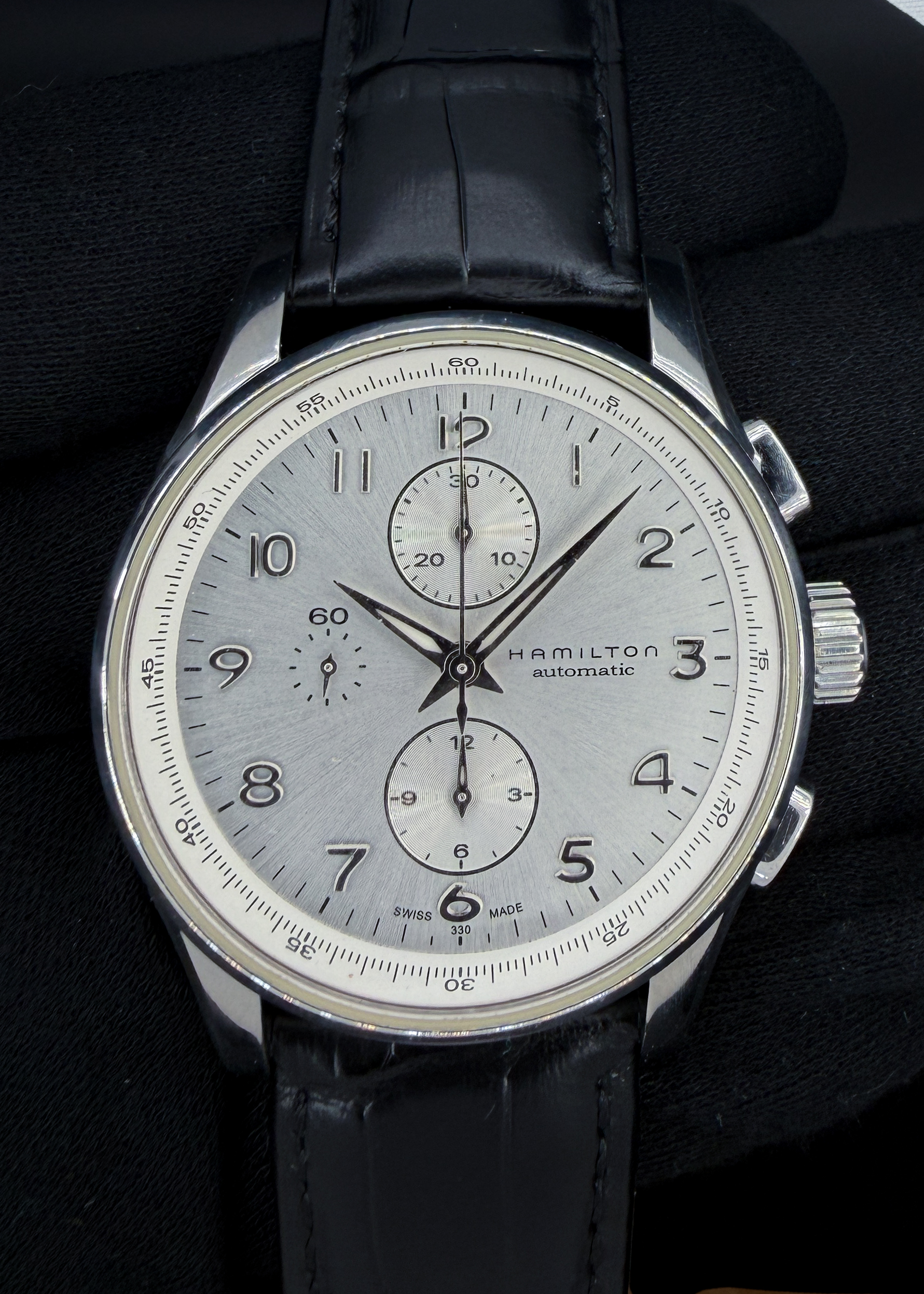 The Hamilton Jazzmaster Maestro H327160 combines classic dress-watch elegance with the substance of a Swiss automatic chronograph. Its 45mm stainless steel case frames a silver sunburst dial with polished applied Arabic numerals, faceted hands, and finely textured chronograph registers. Powered by the respected ETA/Valjoux 7750 automatic movement, it features central chronograph seconds, a 30-minute counter at twelve o’clock, a 12-hour counter at six, and running seconds at nine. A sapphire crystal protects the dial, while the polished bezel and sculpted lugs complement its refined design. Presented on a black leather strap and originally rated for 100 meters of water resistance, the Maestro offers substantial wrist presence with styling suited to both everyday wear and dressier occasions.