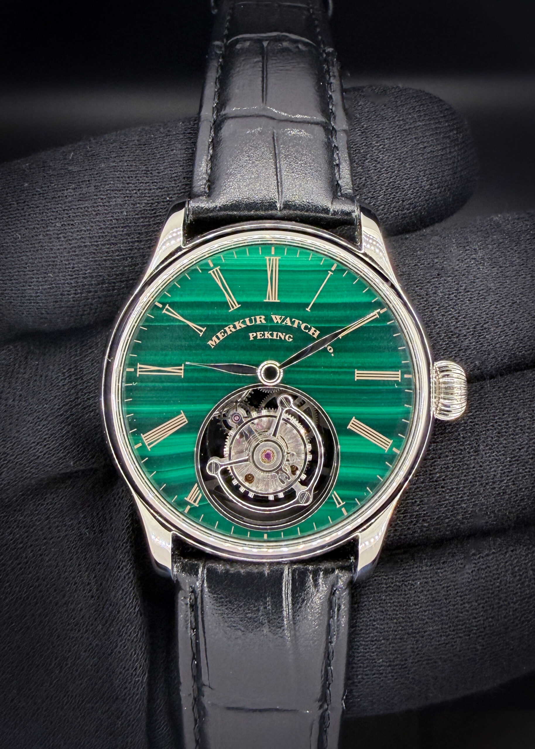 Merkur Watch Co. Peking Tourbillon — Green Malachite Dial — 42mm The Merkur Watch Co. Peking Tourbillon combines a richly banded green malachite dial with gold-tone Roman numerals, polished hands, and a prominently displayed tourbillon at 6 o’clock. The polished stainless steel case measures 42mm in diameter, approximately 12.5mm thick, and 48mm lug-to-lug. Powered by a hand-wound mechanical tourbillon movement, this watch offers a compelling view of its mechanics from both sides. The exhibition caseback reveals the skeletonized bridges and exposed gear train, while a sapphire crystal protects the dial. A black embossed leather strap completes its classic dress-watch presentation. This example is new and includes the travel case, polishing cloth, and instruction book.
