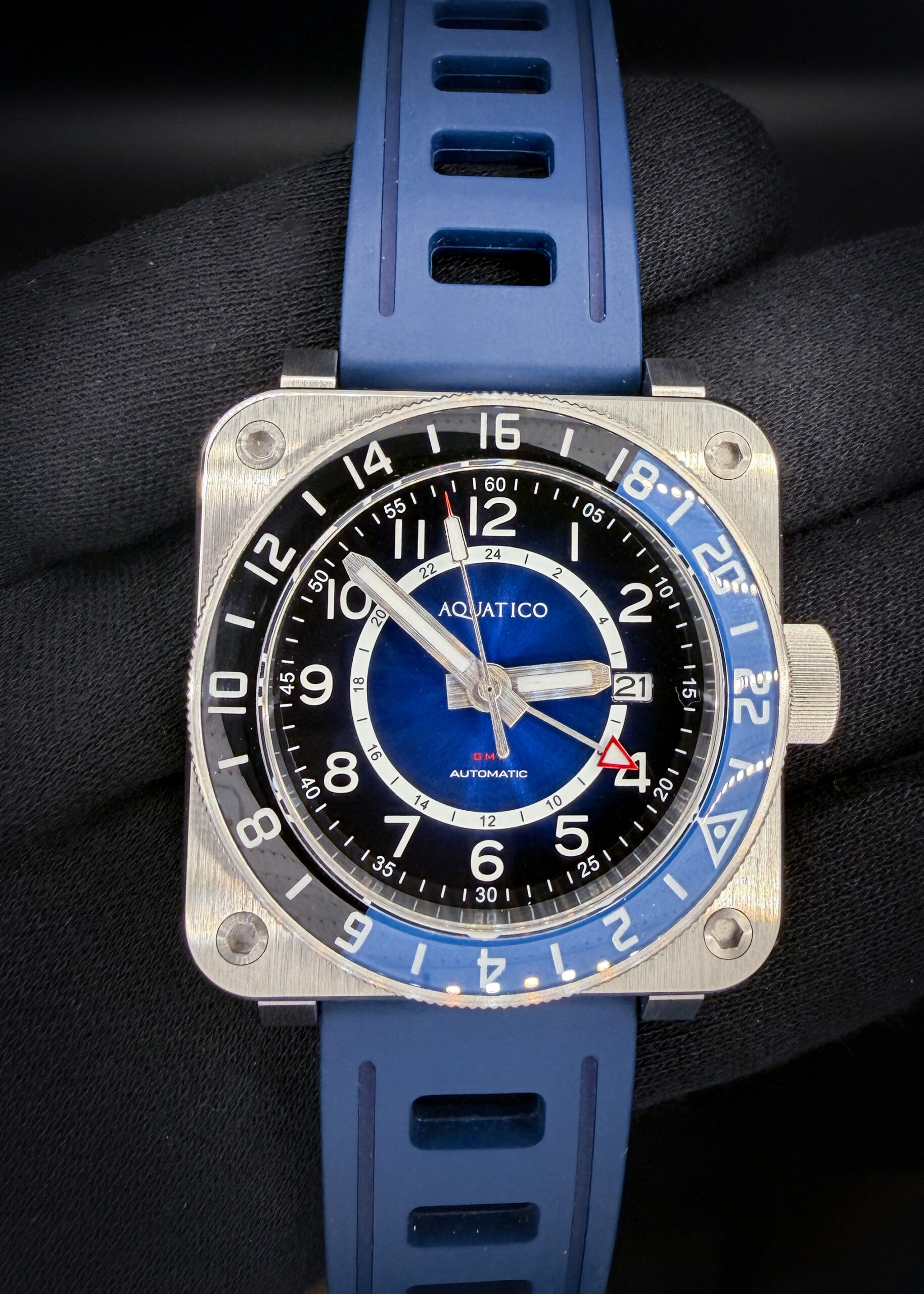 Aquatico Steel Man GMT Blue Automatic — #433/999 — Box & Papers The Aquatico Steel Man GMT pairs a bold square stainless steel case with a blue sunburst dial, contrasting black chapter ring, and blue-and-black 24-hour bezel. Large Arabic numerals, luminous hands, and a red-tipped GMT hand give the watch a distinctive, easy-to-read layout. The case measures 43mm wide, 15.5mm thick, and 50mm lug-to-lug. Inside is a Japanese Seiko NH34A automatic movement with date display and an independently adjustable GMT hand for tracking a second time zone. Sapphire crystal, an exhibition caseback, and a screw-down crown complete the construction. The model carries a manufacturer-rated water resistance of 1,000 meters. Numbered 433/999, this example is presented in new condition and fitted with a blue rubber strap. Includes the outer sleeve, outer box, inner wooden presentation case, instruction card, warranty card dated June 26, 2025, and an additional unworn brown leather strap.
